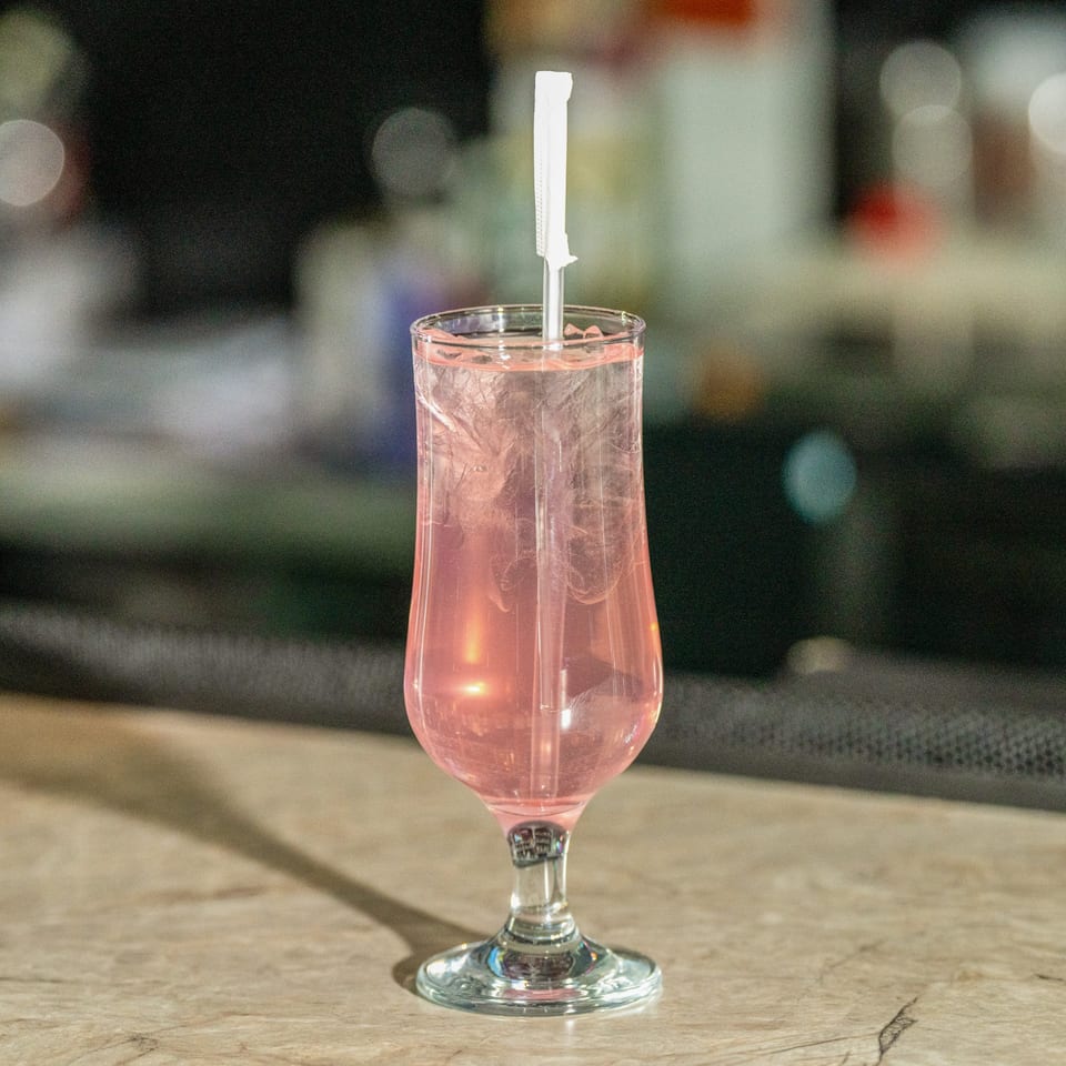 A tall glass of pink drink with ice and a white straw on a wooden table.