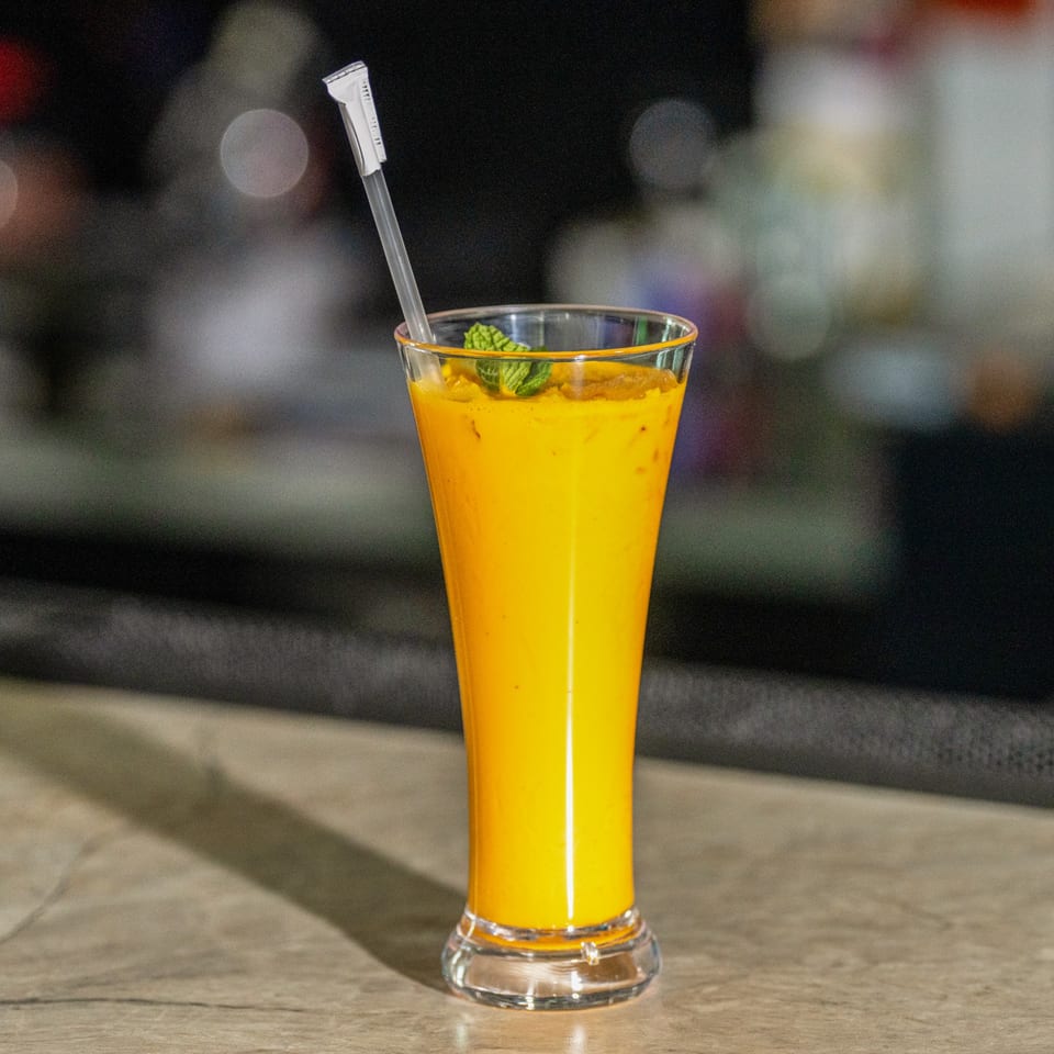 A tall glass of vibrant orange mango lassi, garnished with fresh mint, on a light-colored counter.