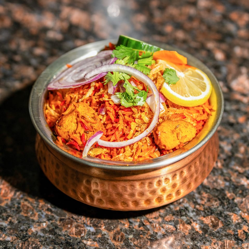 Jhapali Kitchen's Chicken Biryani, a Nepalese dish, served in a copper bowl with lemon, onions, and cilantro.
