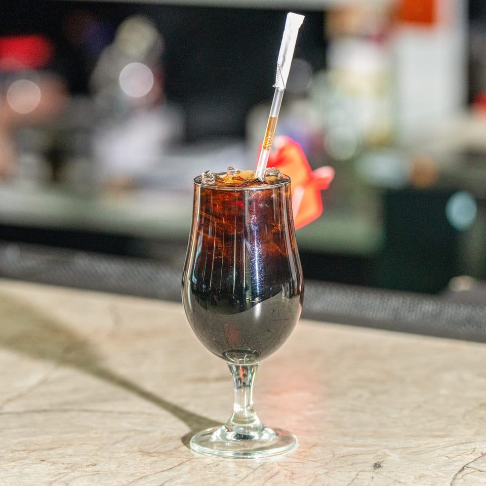 Dark beverage with ice and a straw in a stemmed glass on a light-colored counter.