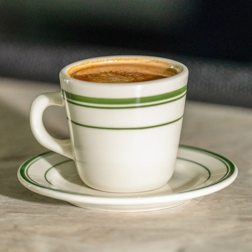 A white cup with green stripes and a matching saucer holds a dark beverage with foam.